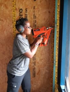 A black lady in a grey shirt and trousers, wearing ear defenders, using a nail gun on a wall.